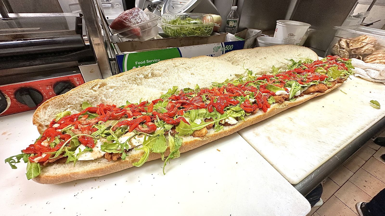A six-foot party sub topped with roasted peppers and arugula being assembled