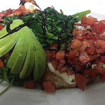 A Plate Of Grilled Chicken Topped With Diced Tomatoes, Sliced Avocado, And Sautéed Greens, Drizzled With Balsamic Glaze.