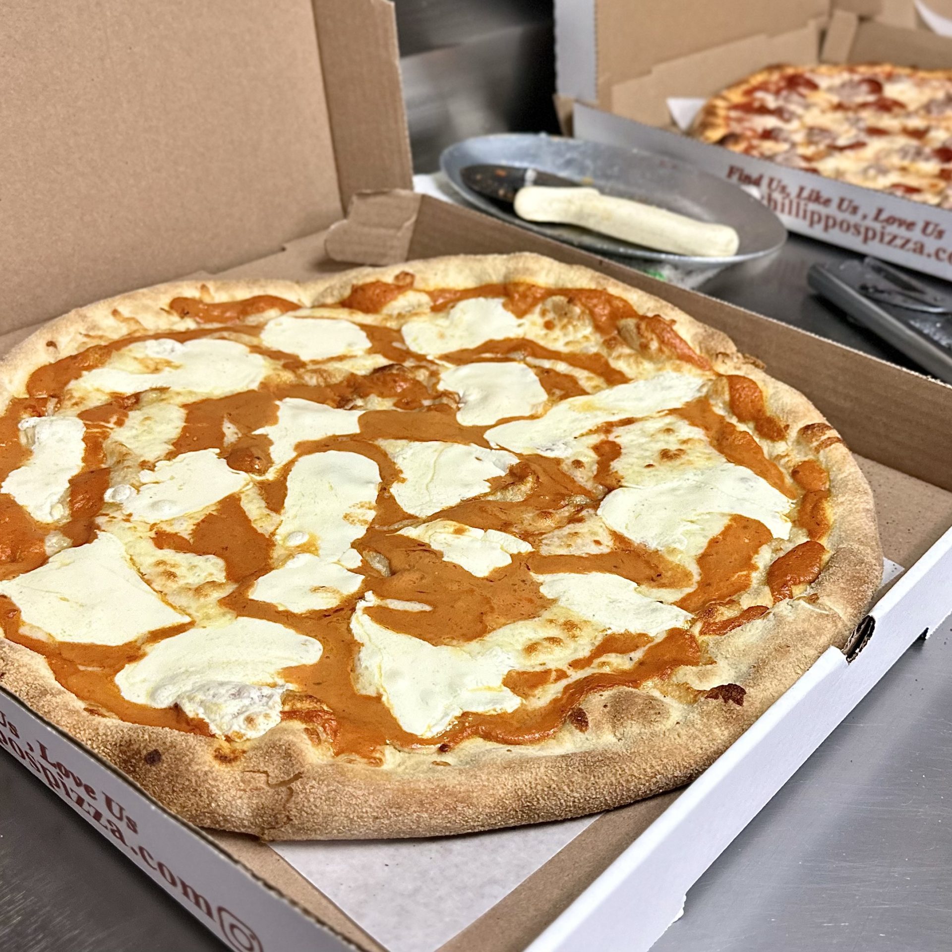 Pizza In An Open Box With Cheese And Sauce, Next To Another Pizza In A Partially Open Box On A Stainless Steel Surface.