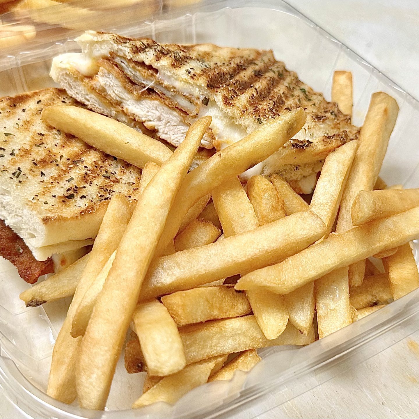 Grilled Sandwich With Cheese And Meat Served In A Plastic Container, Accompanied By A Portion Of French Fries.