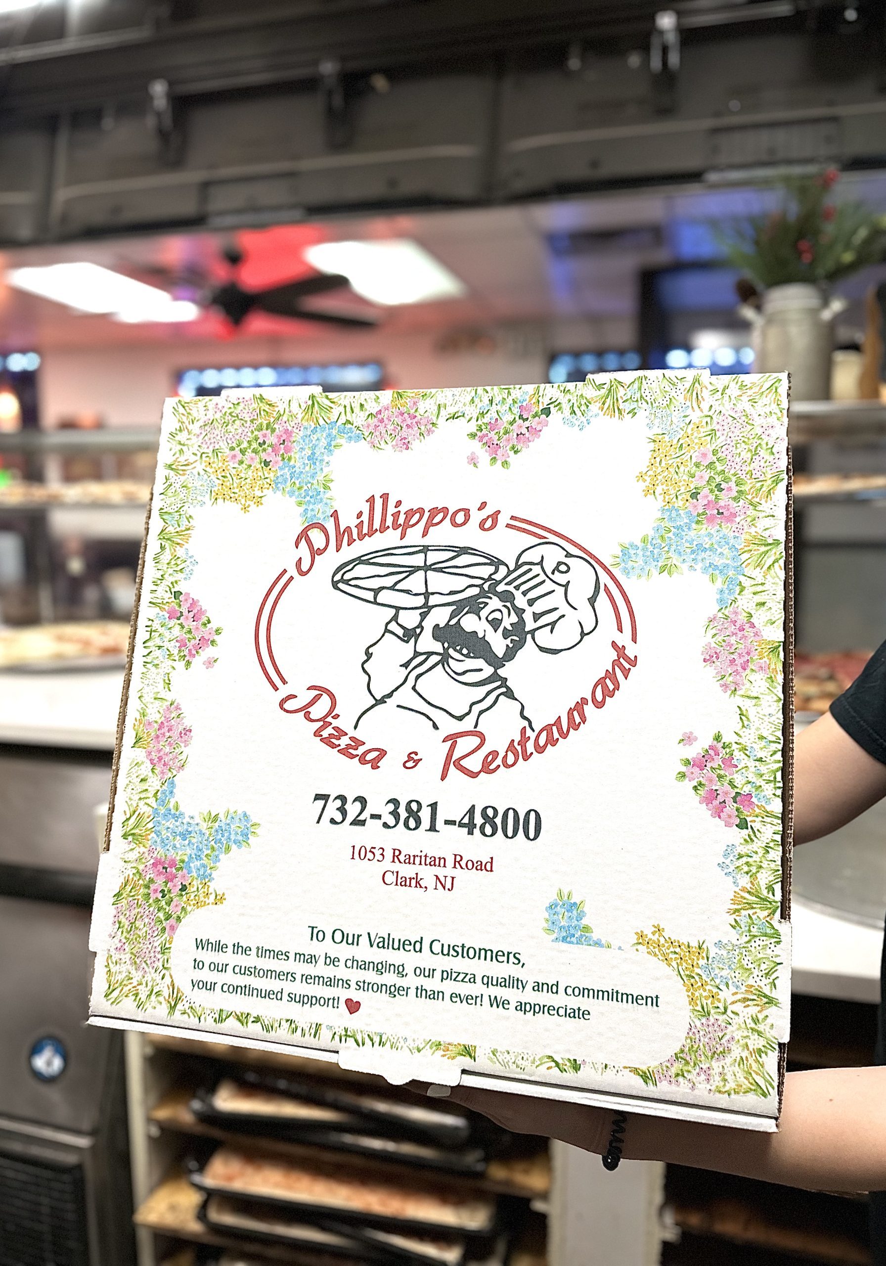 A Person Holds A Phillipo's Pizza & Restaurant Box With A Floral Design And Contact Information In A Kitchen Setting.